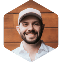 Max, beard and mustache with a hat on, smiling on a background of wood